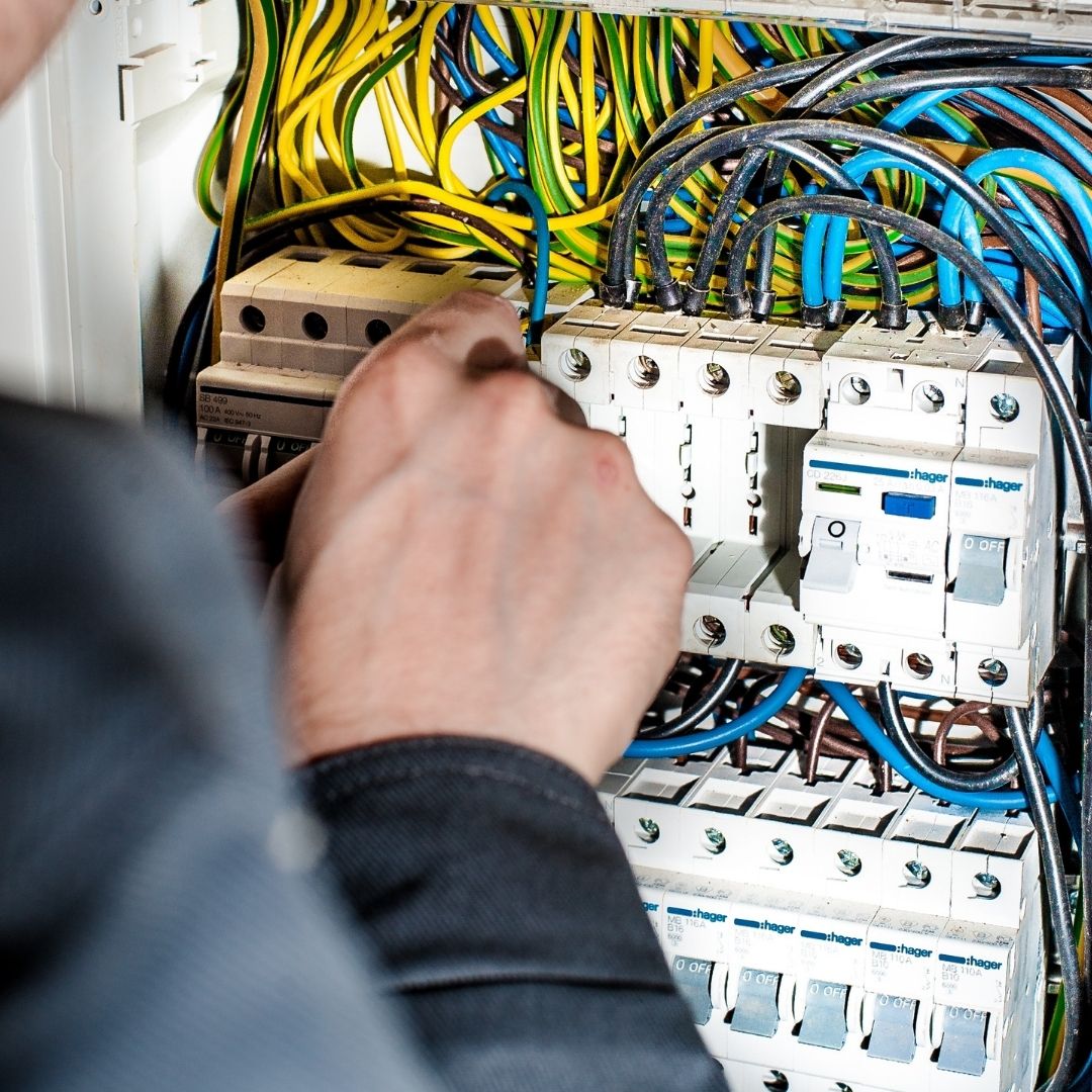A close-up, square-cropped photo of an electrician's hands working inside a complex electrical fuse box. The image shows a dense network of yellow, green, and blue wires connected to several white circuit breakers, with the technician appearing to adjust a connection.